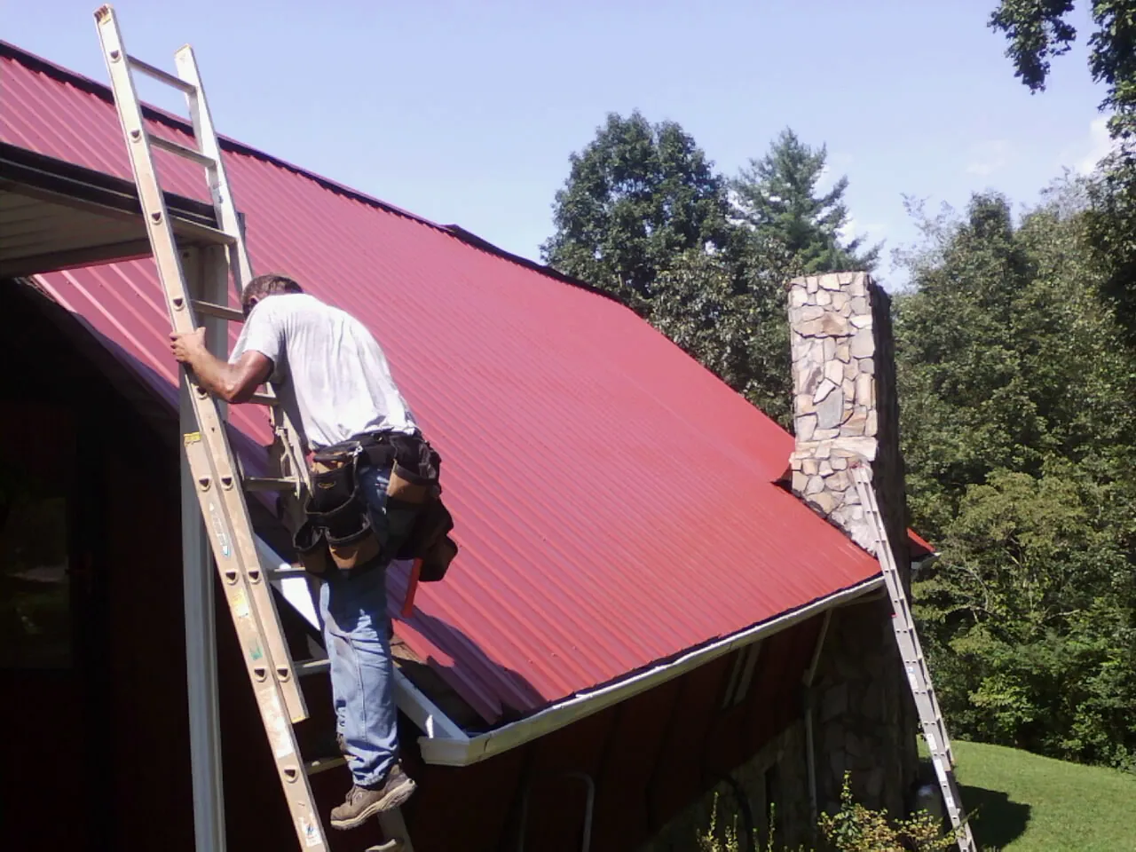 Roofer on a ladder beside a red metal roof and stone chimney — Soffit & Fascia Repair by Roofing Brothers in Glencoe